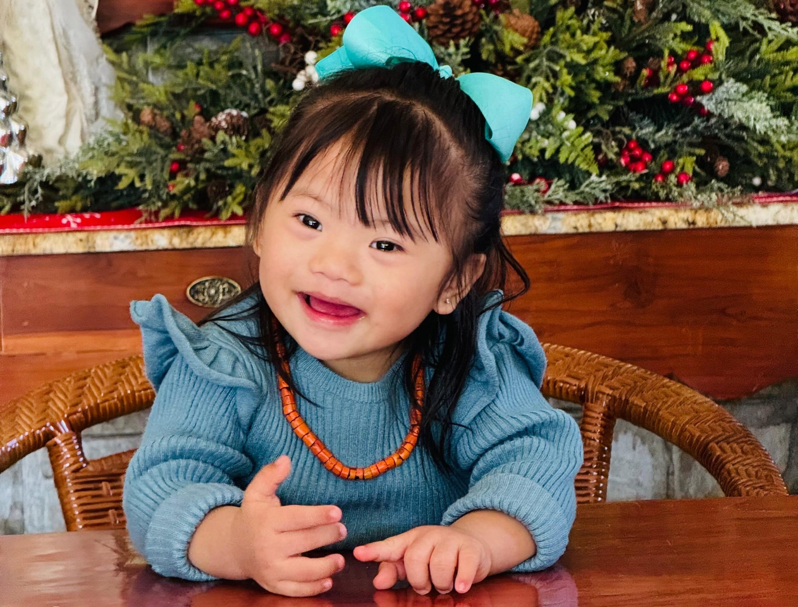 Alashi laughing at a wooden table in a blue knitted jumper and a bright turquoise hair bow
