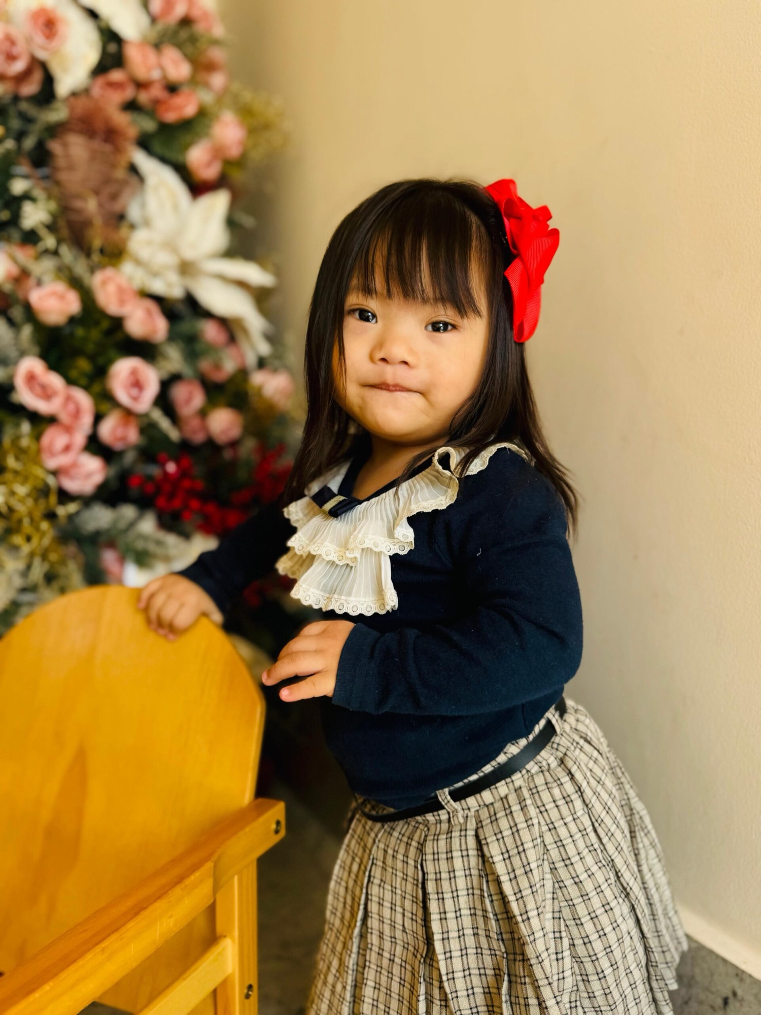 Alashi in a navy dress with a red hair bow, standing beside an arrangement of pink and white flowers
